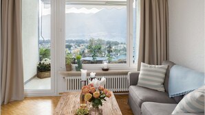 A living room with a view of a city and mountains, a sofa, a coffee table with flowers, and a window with curtains.