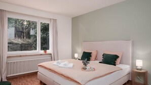 A bedroom with a large bed, two bedside tables, a window with curtains, a radiator, and a view of trees outside. - Lovely apartment for 4 guests with WIFI, TV, balcony and pets allowed (Ascona)