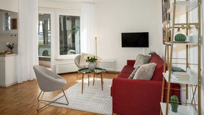 A modern living room with a red sofa, a grey armchair, and a glass coffee table. There is a TV mounted on the wall, a bookshelf, and a window with curtains. - Lovely apartment for 4 guests with WIFI, TV, balcony and pets allowed (Ascona)