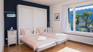 A bedroom with two beds, a large window with a view of mountains, a radiator, and a bedside table with a lamp.