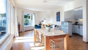 A modern kitchen with white cabinets, a dining area with a wooden table and chairs, and a large window with curtains.