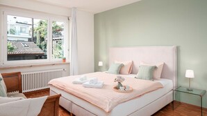Una camera da letto con un letto grande, una finestra che si affaccia su edifici e alberi, un termosifone, una sedia di legno e un tavolino in vetro con una lampada.