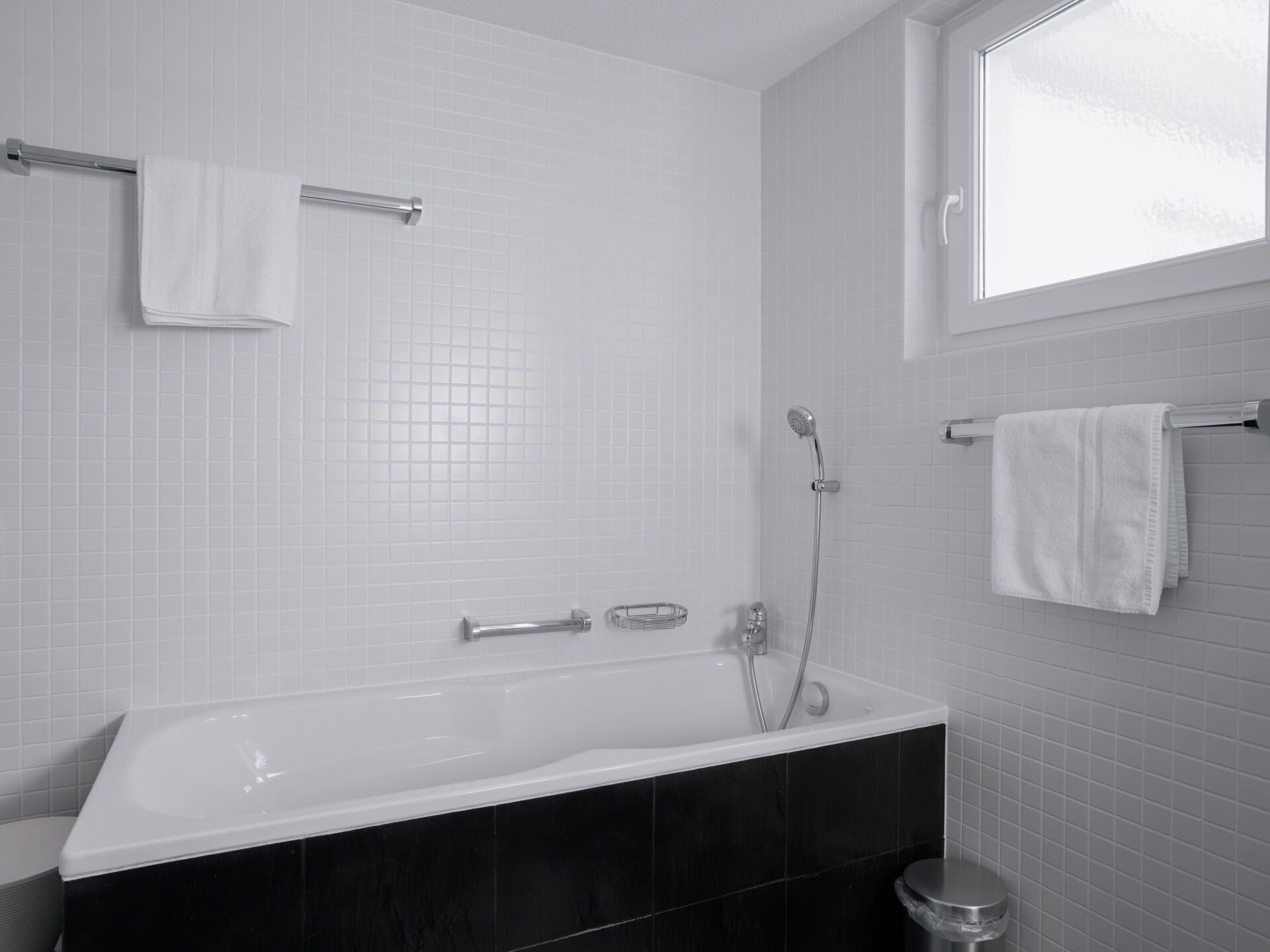 A modern bathroom with a white bathtub, black tiles, a window, and a towel rack.