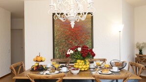A dining room with a wooden table set for breakfast, surrounded by wooden chairs. A chandelier hangs above the table, and a painting of flowers is on the wall behind. - Nice apartment for 4 people with WIFI, TV, balcony and pets allowed (Ascona)