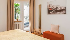A bedroom with a bed, a small orange sofa, a wicker basket, a framed picture on the wall, and a window with curtains. - Nice apartment for 4 people with WIFI, TV, balcony and pets allowed (Ascona)