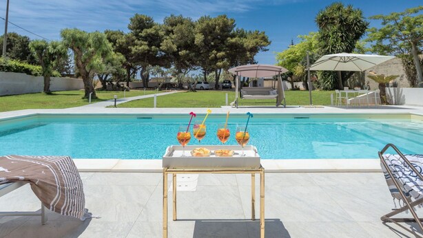 Area piscina con tavolo apparecchiato per bevande e snack, circondata da poltrone prendisole e un pergolato.