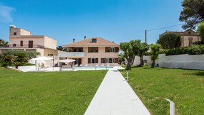 Un vialetto conduce a un edificio con piscina, immerso nel verde e delimitato da un muro bianco.