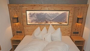 Ein Schlafzimmer mit einem Holzkopfteil, einem großen gerahmten Bild von einer Berglandschaft, einem Bett mit weißer Bettwäsche und zwei Nachttischen mit Lampen.