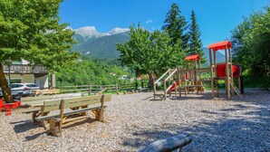 Un'area giochi con panchine di legno e uno scivolo, circondata da alberi e montagne sullo sfondo.
