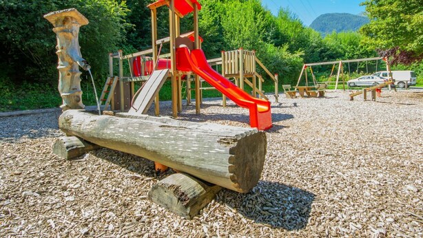 Un’area giochi con uno scivolo di legno e strutture per arrampicarsi, circondata da alberi e un fondo in ghiaia.