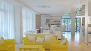 Et lyst, moderne loungeområde med gule sofaer og hvide borde, store vinduer med lette gardiner samt et synligt skilt med udgang.