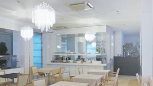 Et moderne spiseområde med hvide borde og stole, et køkken i baggrunden og dekorative lysekroner.