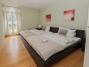 A double bed with a wooden headboard, a bedside table with a lamp, and a view of a balcony through a window with curtains.