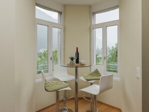 Habitación de esquina con dos ventanas, una mesa de vidrio y cuatro sillas blancas con cojines verdes.