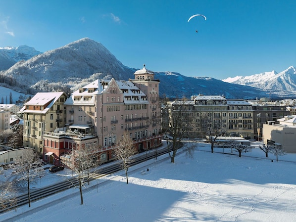 Un área urbana cubierta de nieve con edificios históricos, una vía de tren y montañas al fondo.