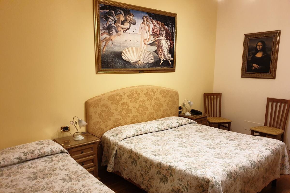 A hotel room with two beds, a painting of a classical scene, a framed portrait, and a wooden chair.