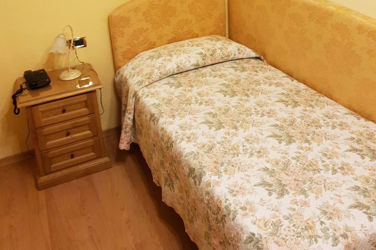 A bedroom with a bed, a nightstand with a lamp, a telephone, and a painting on the wall.