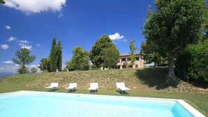 Una piscina con cuatro sillas reclinables blancas, rodeada de césped y árboles, y un edificio al fondo.