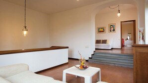 Amplia sala de estar con techo alto, paredes blancas y un banco de madera. Hay un sofá blanco, una mesita pequeña con un adorno decorativo y una escalera que conduce a un dormitorio.