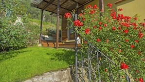 Une maison en bois avec un toit de chaume, entourée d’un jardin bien entretenu où poussent des roses rouges, et un chemin de pierres qui mène à l’entrée.