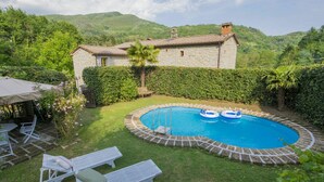 Une maison en pierre avec une piscine, entourée d’une bordure en briques, deux chaises longues et un banc.
