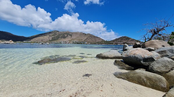 Una playa con agua cristalina, rocas grandes y una montaña al fondo.