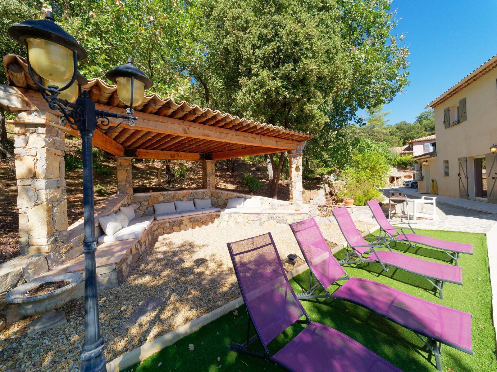 Un espace extérieur aménagé, abrité et doté de murs en pierre et d’une pergola, entouré de verdure et bordé d’un bâtiment au toit recouvert de tuiles.