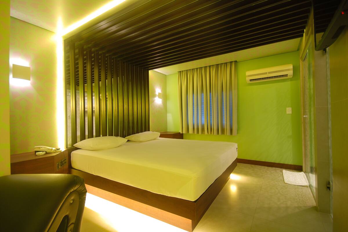 A hotel room with a bed, a nightstand, a chair, a window with curtains, and an air conditioning unit.