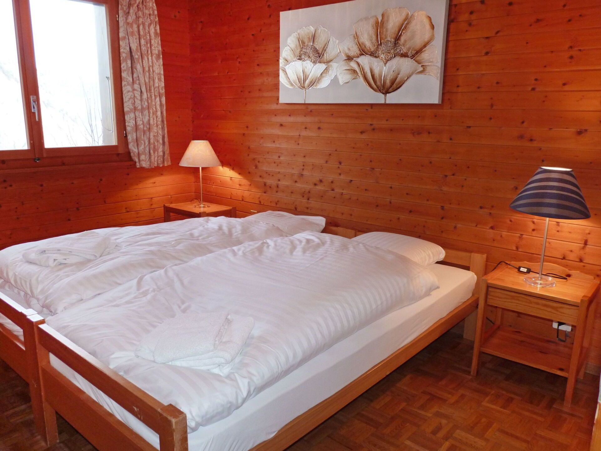 A wooden-paneled room with a large bed, two bedside tables, a window with curtains, and a painting of flowers on the wall.