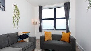 Een moderne woonkamer met twee grijze banken, waarvan er één een geometrisch kussen heeft en de andere twee oranje kussens. Op een houten tafel in het midden staat een groene vaas.