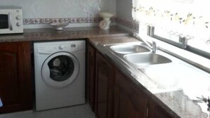 A kitchen with wooden cabinets, a washing machine, a microwave, and a sink. - Apartamentos Odeceixe (Aljezur)