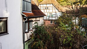 Une maison traditionnelle à colombages, avec une façade blanche et une porte verte, entourée d’un feuillage dense et se dressant sur un terrain vallonné.