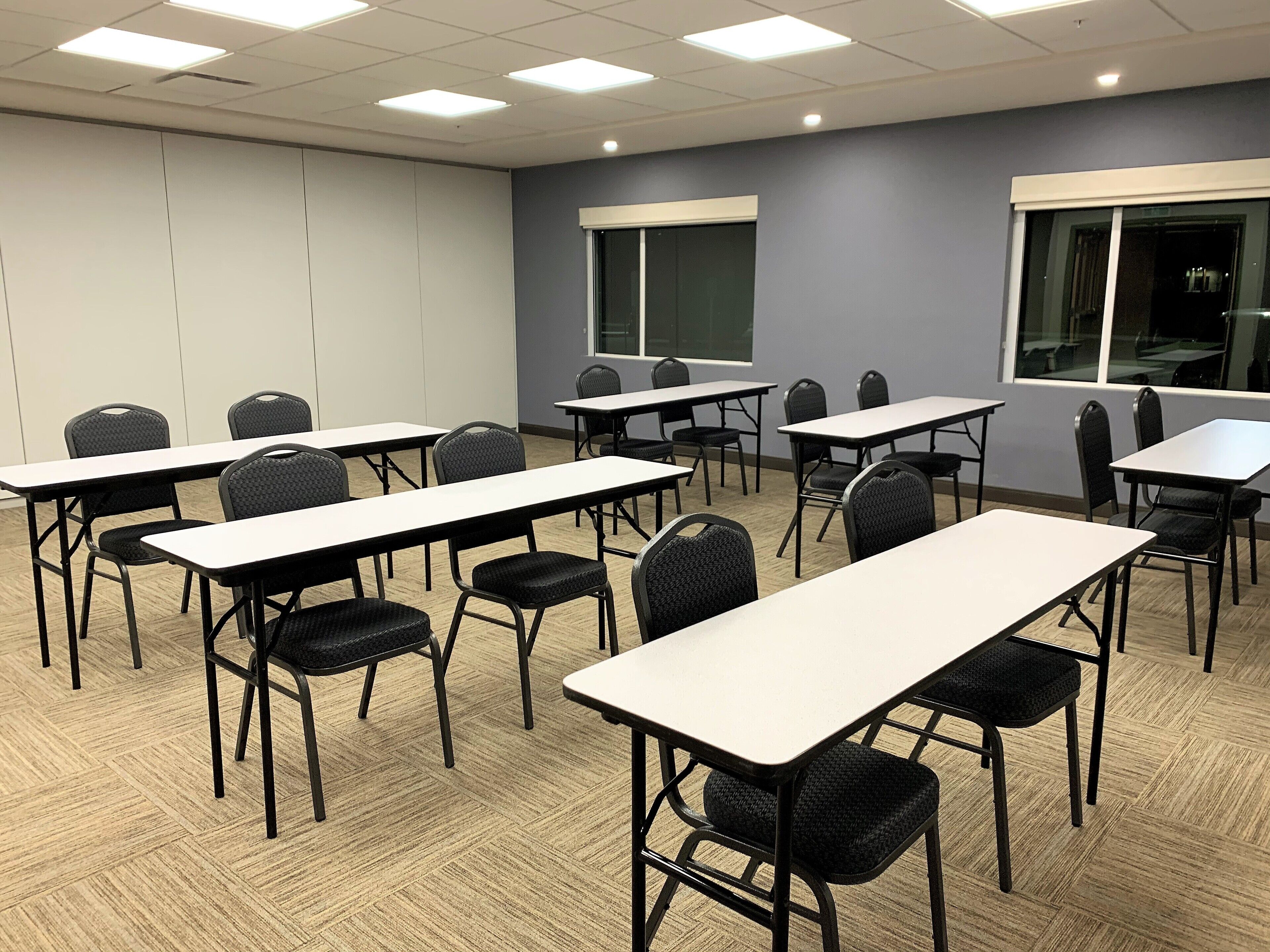 Un salón de conferencias con varias mesas y sillas, ventanas amplias y un suelo con estampado tipo espiga.