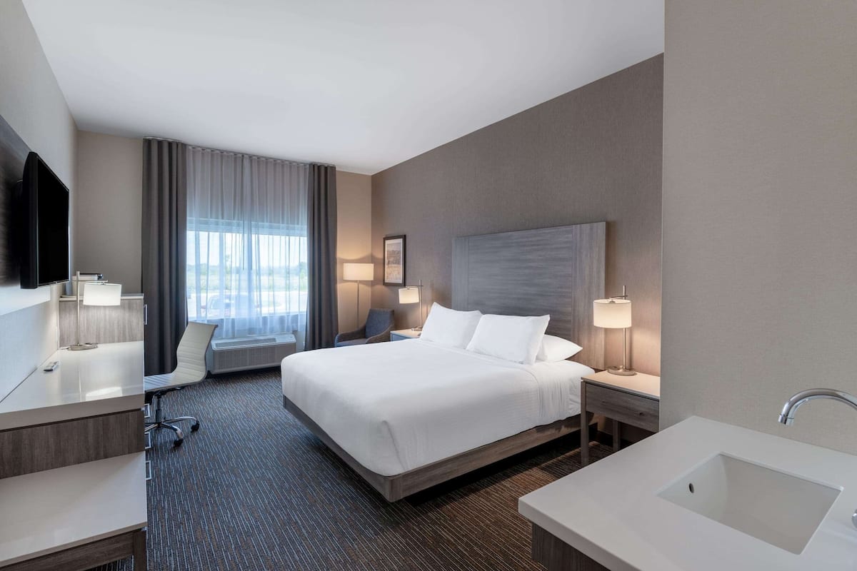 A hotel room with a large bed, a desk, a chair, a desk lamp, a bedside table with a lamp, a large window with curtains, and a bathroom with a sink and mirror.