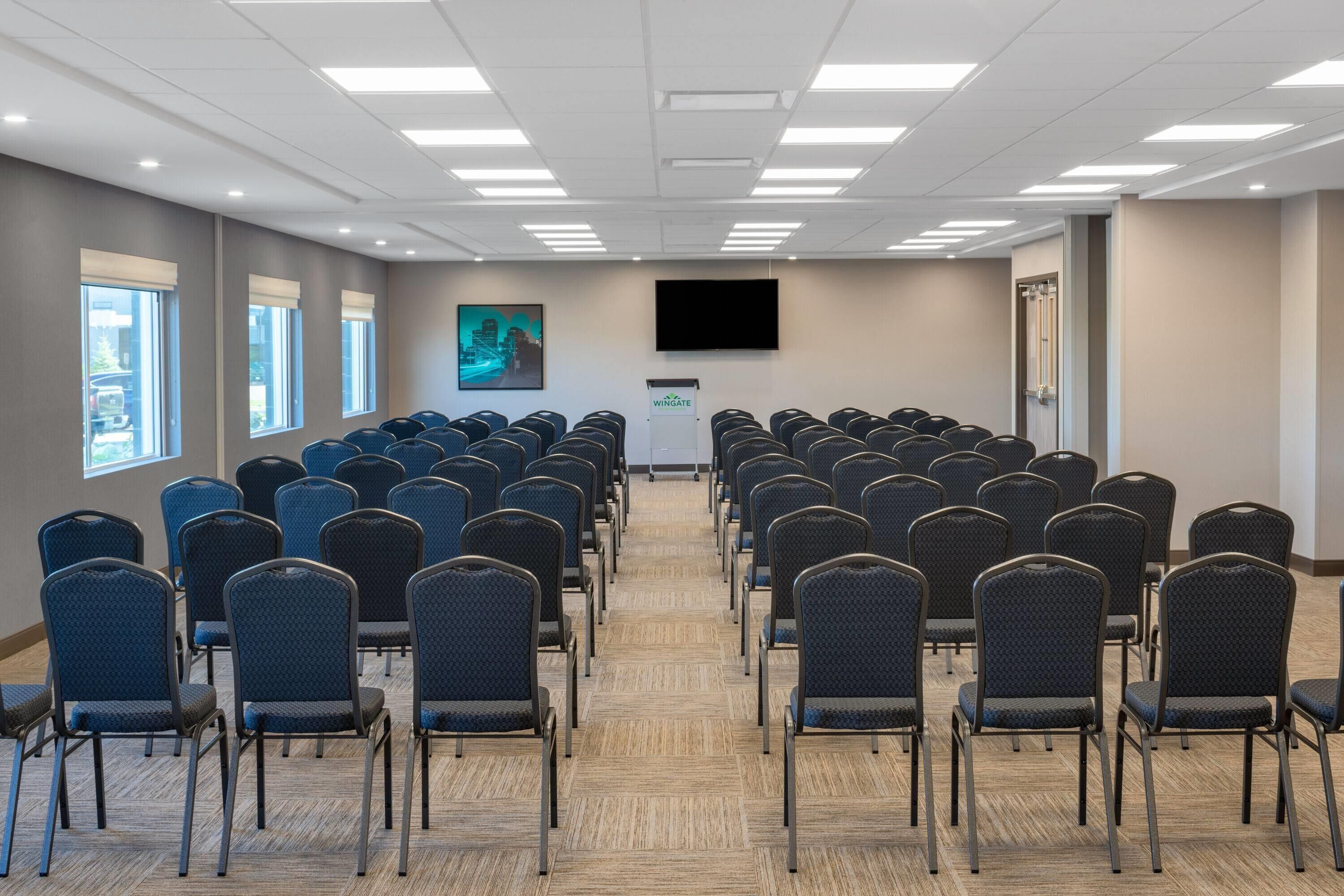 A conference room with rows of chairs, a podium, a large screen, and windows with blinds.