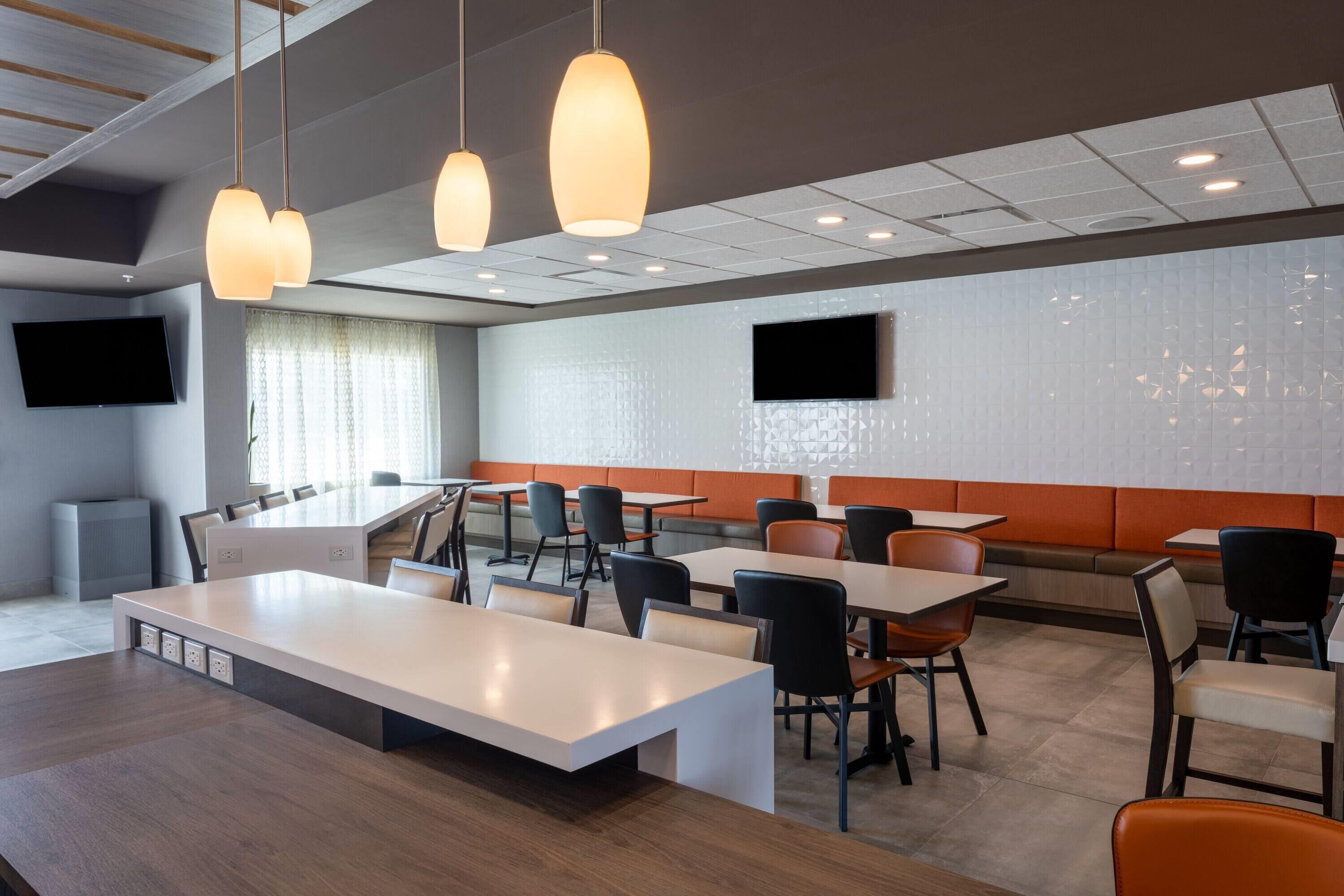 A modern dining area with tables and chairs, pendant lights, a flat-screen TV mounted on the wall, and a seating area with a cushioned bench.