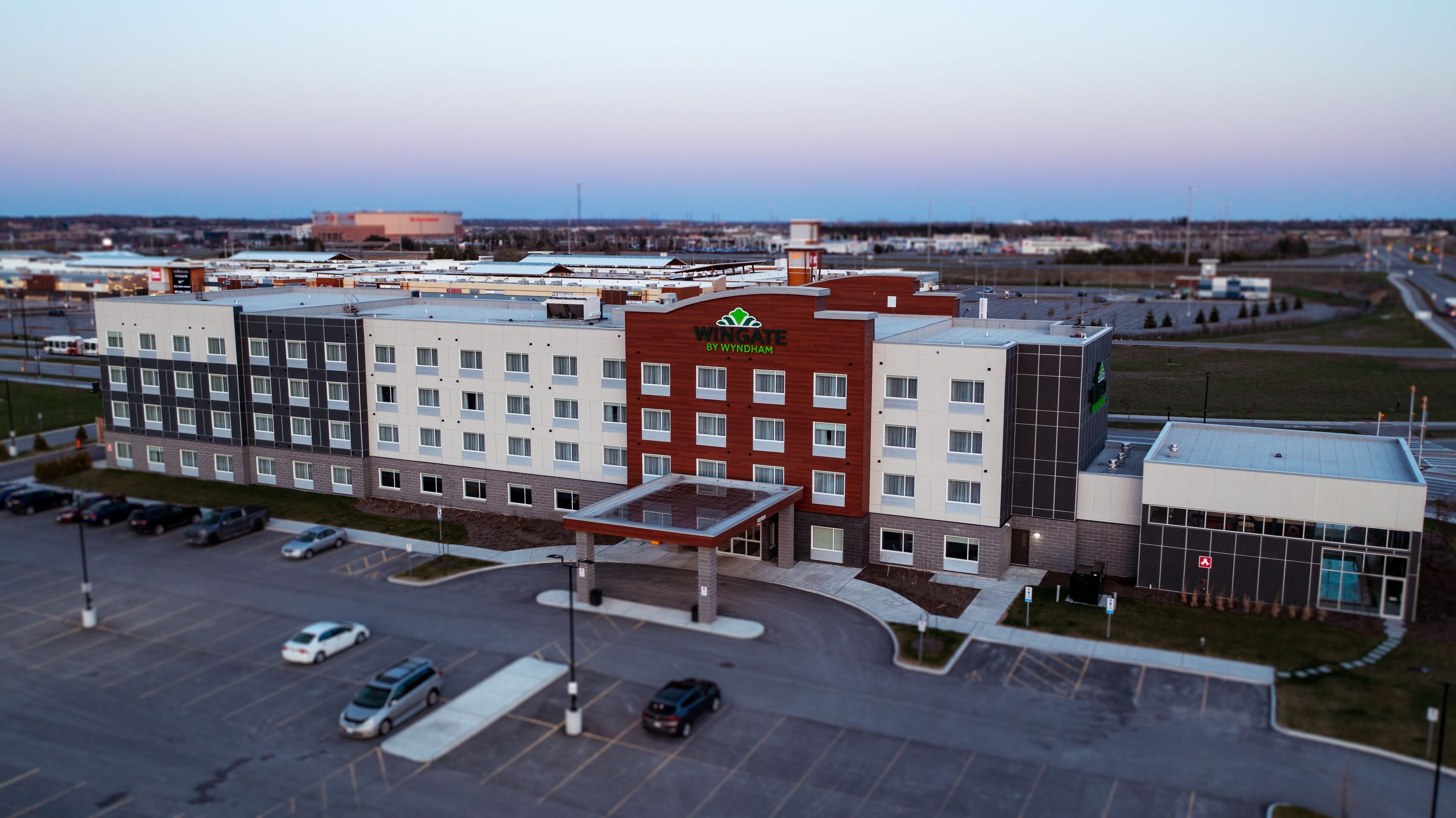 Un hotel de varios pisos con un llamativo letrero de 'Wingate by Wyndham', un estacionamiento con varios autos y una vista despejada del entorno.