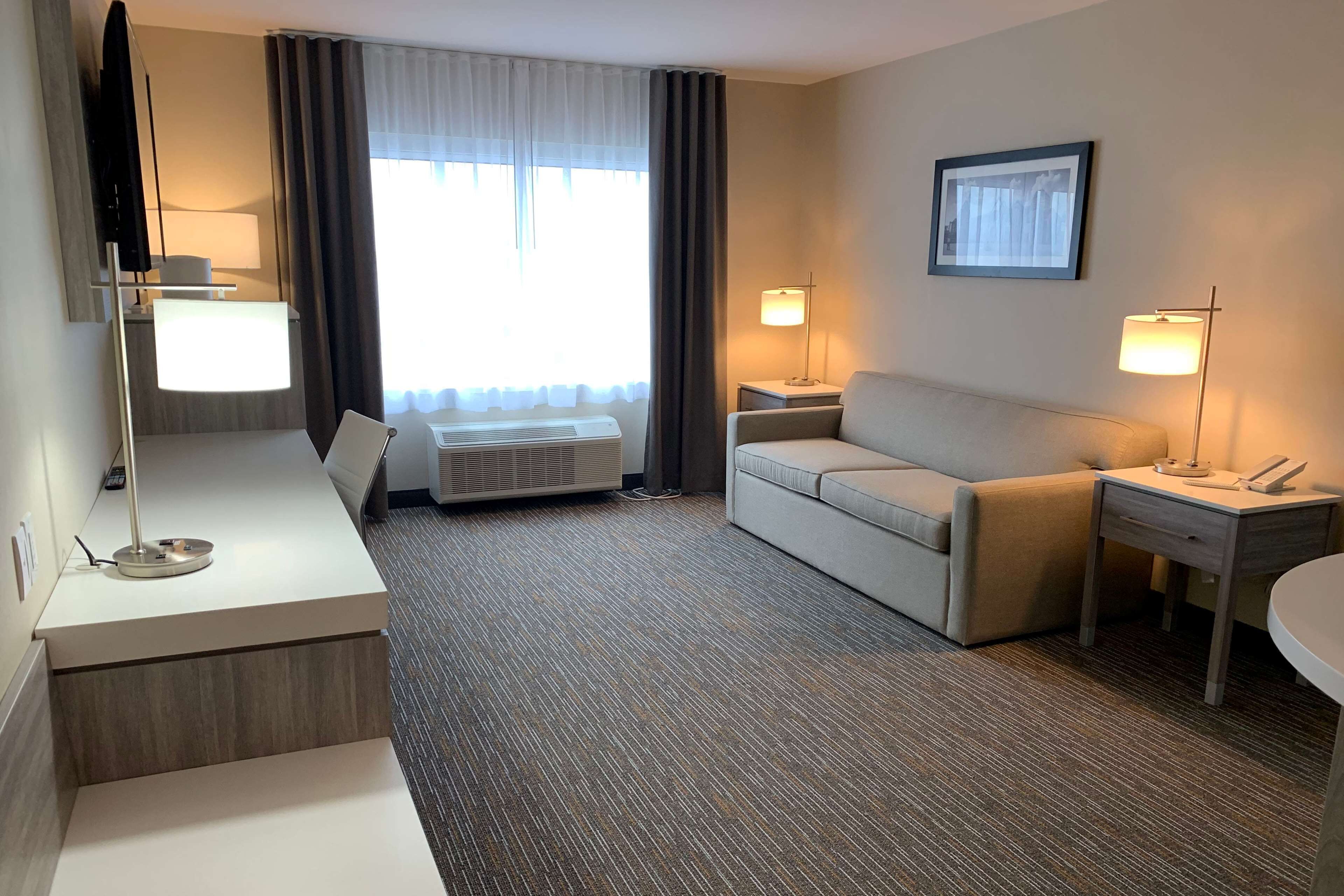 Une chambre d’hôtel comprenant un lit, un bureau avec une lampe, un canapé, une table de chevet et une fenêtre avec des rideaux.