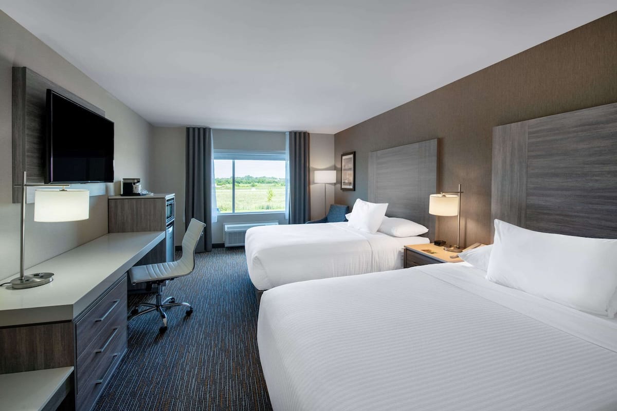 A hotel room with two beds, a desk with a chair, a TV, and a window with a view of greenery.