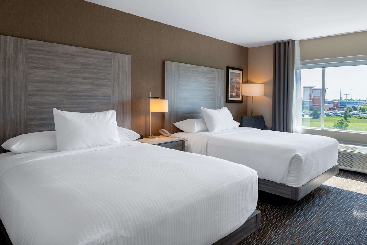 A hotel room with two beds, a large window with a view of a parking lot, and a modern design with neutral colors.