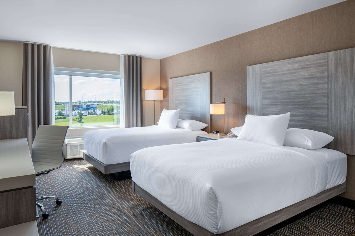 A hotel room with two beds, a desk, and a chair. There is a large window providing a view of the outdoors.