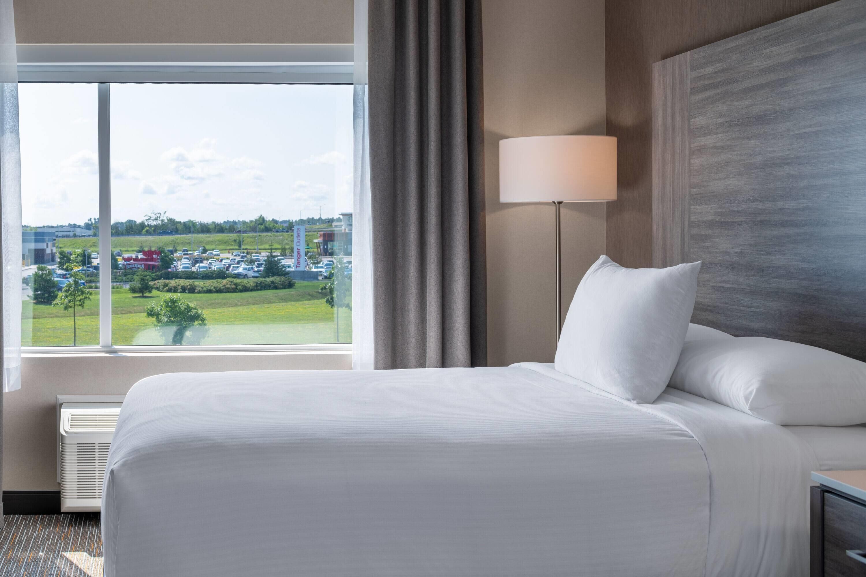 Une chambre d’hôtel avec un grand lit, une vue sur un parking, une lampe et une tête de lit fixée au mur.