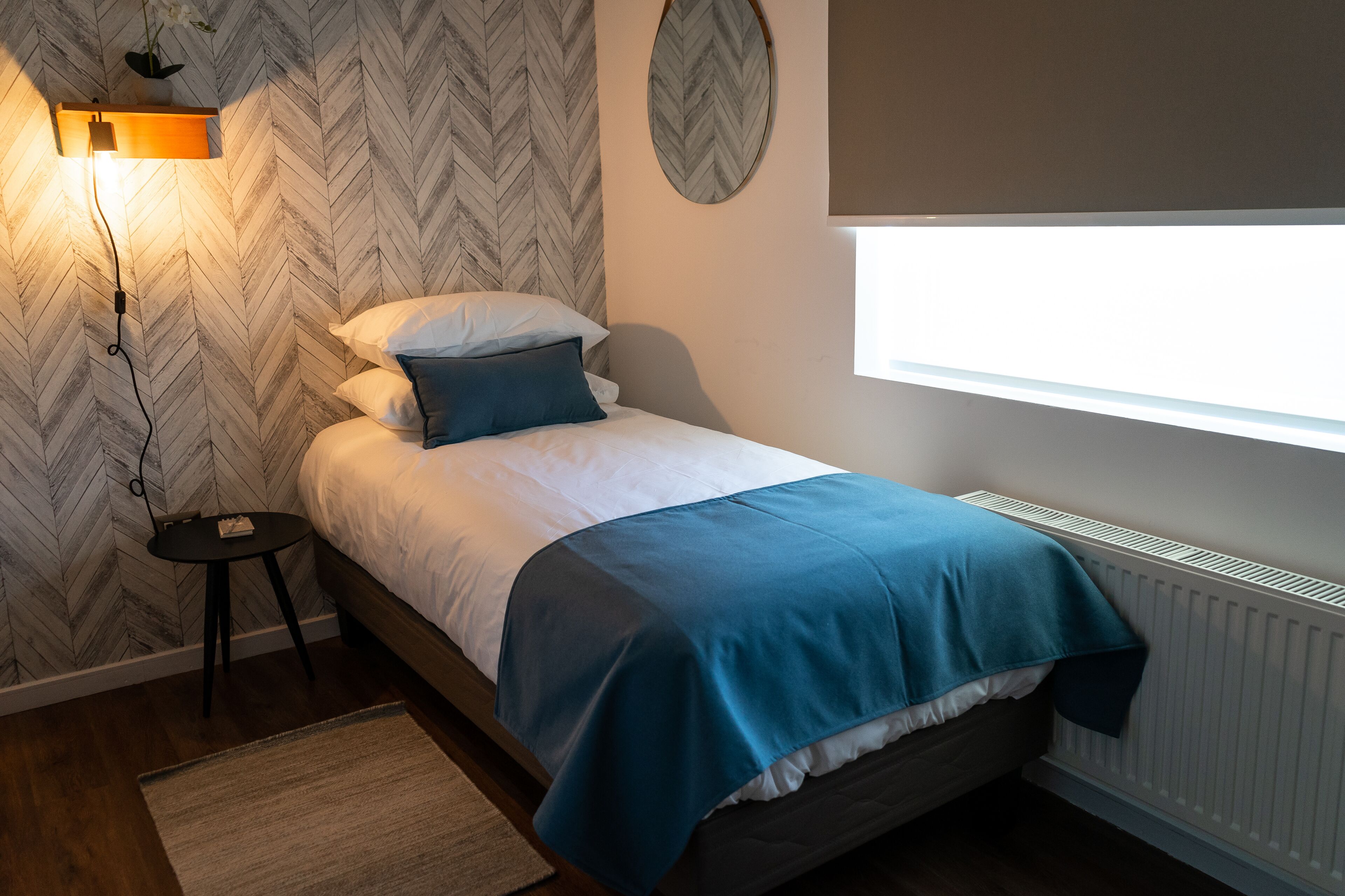 Una cama bien hecha con una colcha azul y blanca, una mesita de noche con una lámpara y un papel tapiz con estampado de espiga.