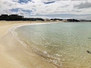 A beach with clear water, sandy shore, and buildings in the background.
