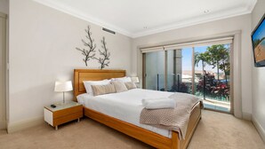 Un dormitorio con cama de madera, ropa de cama blanca, una mesita de noche con lámpara y vistas a un balcón con mobiliario exterior.