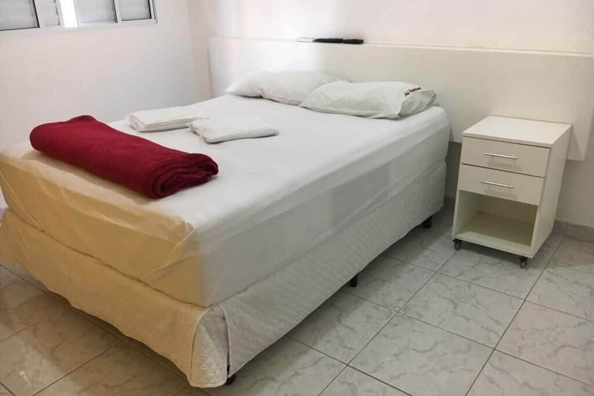 Un lit bien fait, avec une serviette rouge, un oreiller blanc et une taie d’oreiller blanche ornée d’un motif. On aperçoit une petite table de chevet avec des tiroirs et une lampe fixée au mur.