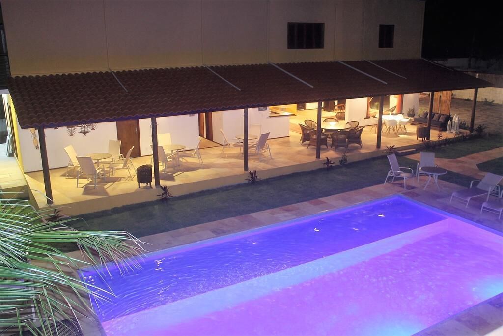 Uma área de estar coberta ao ar livre, com mesas e cadeiras, uma piscina e um edifício com telhado ladrilhado.