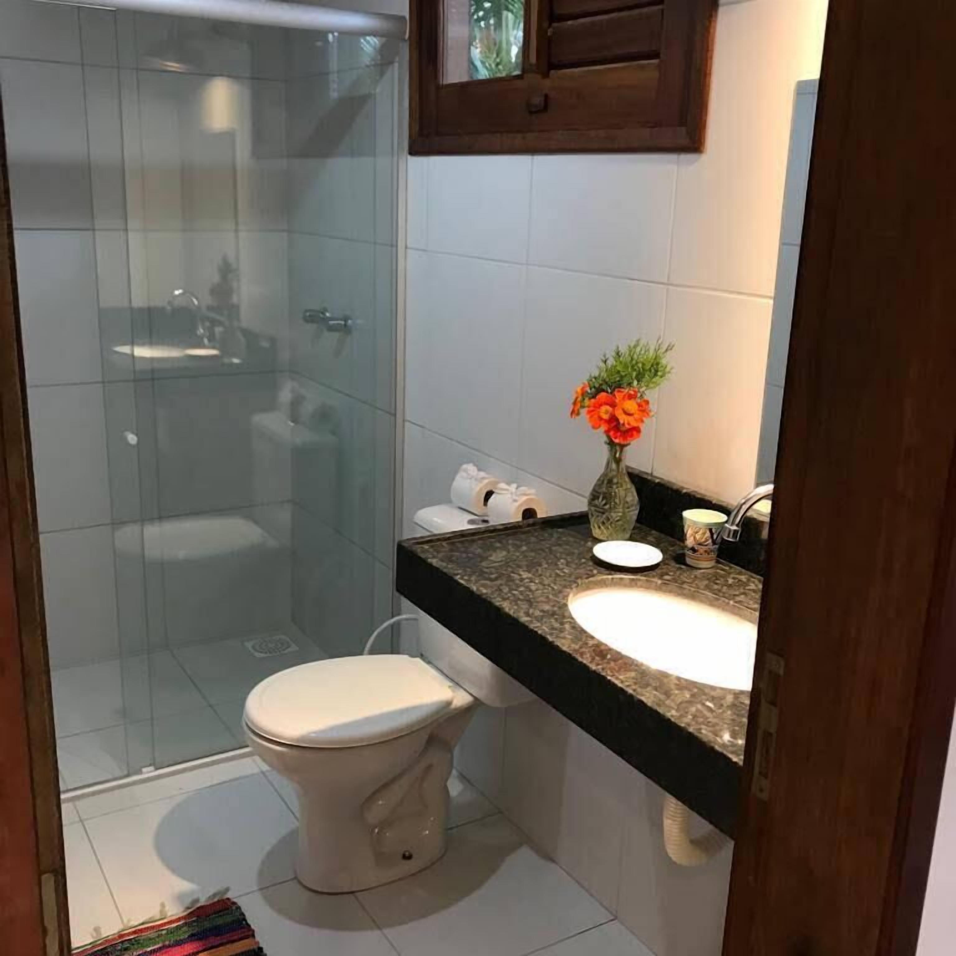 Un baño con cabina de ducha de vidrio, inodoro, lavamanos y un florero con flores.