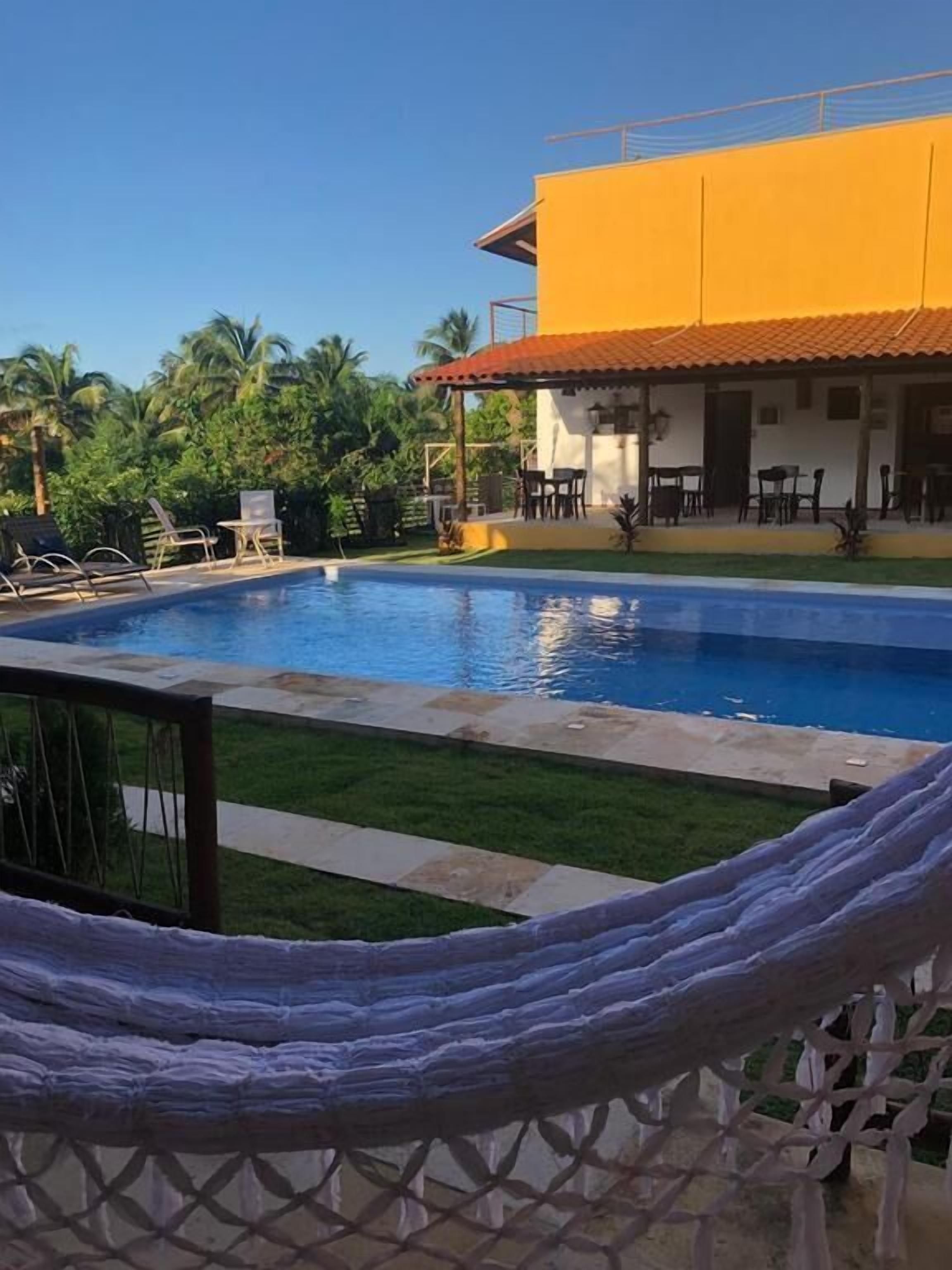 Un área de piscina con sillas de descanso y una hamaca, rodeada por un edificio con techo de tejas y un patio cercado.