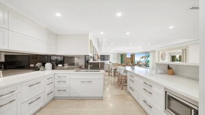 A modern kitchen with white cabinetry, a central island, and a dining area with wicker chairs. - Unit 41 -  3 Bed Garden View (TERRIGAL)